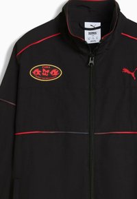 Svart jacka med röda accenter, med dragkedja fram, hög krage och ett broderat Sonic the Hedgehog-logga. Slät textur.