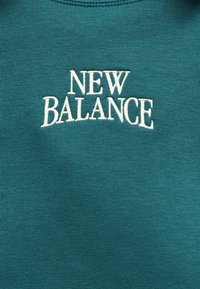 Λευκό κεντημένο κείμενο "NEW BALANCE" σε φόντο τιρκουάζ πλεκτού υφάσματος, κεντραρισμένο και με κεφαλαία γράμματα.