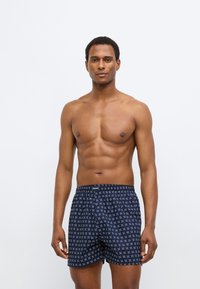 Μπλε ναυτικό βαμβακερά μποξεράκια με γεωμετρικό λευκό σχέδιο, ελαστική μέση και άνετη εφαρμογή, παρουσιασμένα σε αρσενικό μοντέλο.