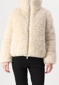 Flauschige cremefarbene Jacke mit Reißverschluss, hohem Kragen, elastischen Bündchen und Saum. Weiche Textur mit flauschigem Finish, kombiniert mit einer schwarzen Hose.