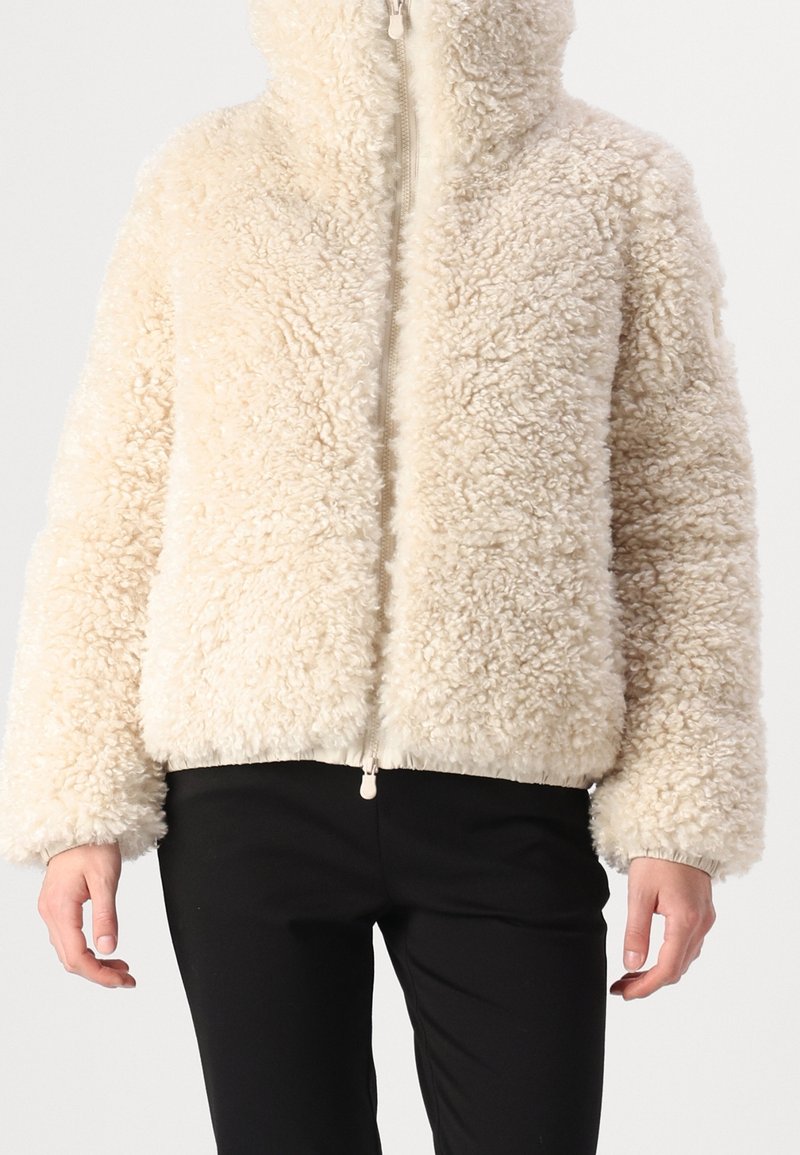 Flauschige cremefarbene Jacke mit Reißverschluss, hohem Kragen, elastischen Bündchen und Saum. Weiche Textur mit flauschigem Finish, kombiniert mit einer schwarzen Hose.