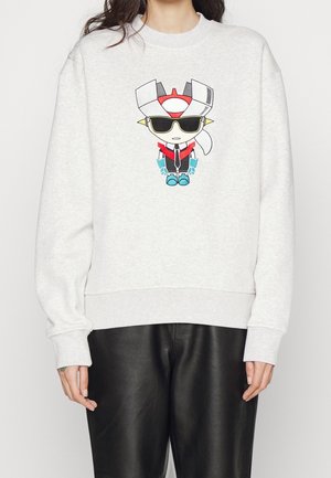 Person iført en lysegrå sweatshirt med en tegneserie-robot iført solbriller trykt på brystet, kombineret med sorte læderbukser.