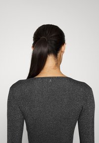 Femme aux longs cheveux bruns attachés en queue de cheval, portant un haut à manches longues gris foncé à paillettes, vue de dos sur un fond clair uni.