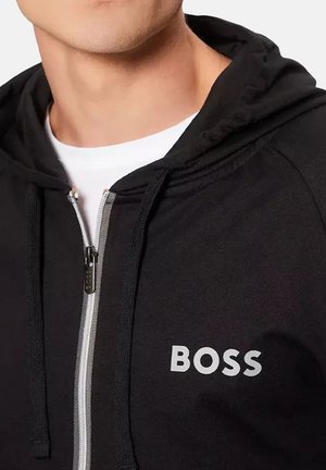 Hombre con sudadera negra con cremallera y el texto "BOSS" impreso en el pecho, sobre una camiseta blanca, mostrando el cuello y la parte inferior de la cara.