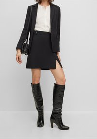 Czarny dopasowany żakiet na białej bluzce, w połączeniu z dopasowaną czarną spódnicą z bocznym rozporkiem. Czarne skórzane kozaki do kolan o błyszczącej powierzchni.