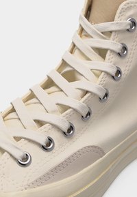 Κοντινό πλάνο λευκού καμβαδένιου αθλητικού παπουτσιού με λευκά κορδόνια περασμένα από ασημένια οπές και μπεζ καουτσούκ σόλα με ραμμένη λεπτομέρεια.