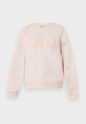 Femme portant un sweat-shirt rose clair, un jean bleu à jambes larges, des chaussures blanches pointues et un sac à épaule marron foncé texturé.