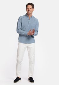 Chemise bleu clair à boutons avec une poche, pantalon blanc et chaussures noires à enfiler. Tenue décontractée présentée sur un fond neutre.
