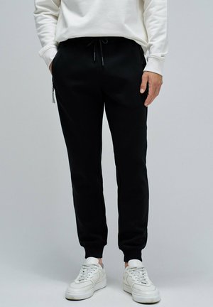 Pantalones de chándal negros hechos de tela suave, con una cinturilla elástica con cordón, bolsillos laterales y puños acanalados. Combinados con zapatillas blancas.