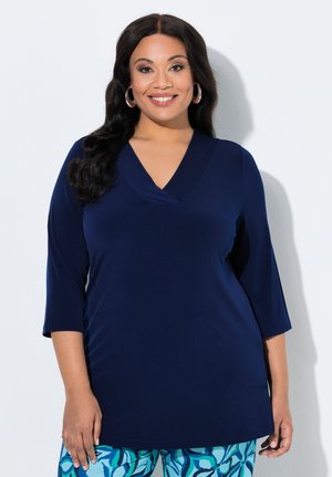 Usměvavá žena s dlouhými tmavými vlasy má na sobě tmavě modrý top s výstřihem do V a tříčtvrtečními rukávy a modré vzorované kalhoty, stojící před bílou zdí.