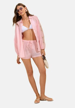 Kvinde iført hvid bikinitop, løs lyserød skjorte, matchende shorts, solbriller, holder en lille taske, står i beige sandaler.