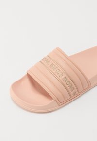 Rosa gummisandale med tre pregete horisontale stropper som har merkevarenavnet i gull. Glatt tekstur og åpen tå-design.