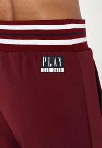 Bordowe spodnie dresowe z białym i granatowym paskiem na talii. Zawierają metkę z napisem "PLAY" oraz "EST. 2006" w kontrastującym wzorze.