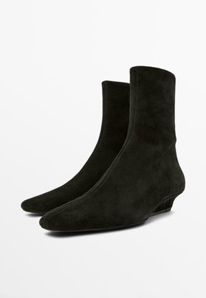 Pár černých semišových kotníkových bot s nízkými podpatky ve tvaru koťátka a špičatými špičkami, zobrazených vedle sebe na bílém pozadí.