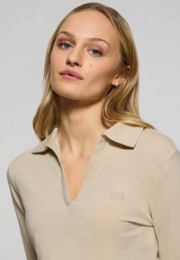 Beige långärmad polo med V-ringning och krage. Har en diskret "POLO CLUB"-logotyp i en ljus textur på bröstet.