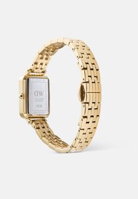 Montre-bracelet en acier inoxydable doré avec un cadran rectangulaire, un bracelet à maillons et une finition polie. Présente un fond blanc avec une marque et un numéro de série.