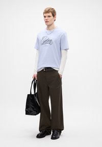 Jasnoniebieska koszulka bawełniana z czarnym logo w formie pisma, nałożona na białą koszulę z długim rękawem; zestawiona z oliwkowymi spodniami o szerokich nogawkach i czarnymi butami.