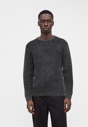 Homme portant un pull en tricot texturé gris foncé et un pantalon noir, debout devant un fond clair uni.