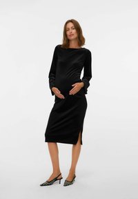 Vestido de maternidad de terciopelo negro con mangas largas y aberturas laterales. Modelo llevando tacones de patrón blanco y negro. Textura suave, diseño ajustado.