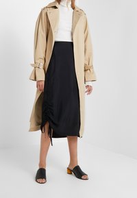 Beige trenchcoat med rullade ärmar, svart satin kjol med infällda detaljer, och svarta öppna sandaler med träklack.
