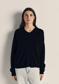 Črn, gumbast cardigan s okroglo ovratnikom in rebrastimi manšetami. Mehka struktura, udoben kroj, se dobro ujema s svetlimi kavbojkami z visokim pasom.