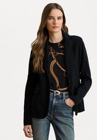 Zwarte blazer met een tailored ontwerp, met schouderflappen en voorkwamen, gedragen over een met patroon bedekte top, in combinatie met vervaagde blauwe jeans.