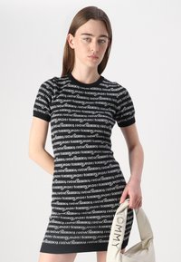 Robe noire à manches courtes avec un motif répétitif du texte "tommy jeans" en blanc, silhouette ajustée, col côtelé, et un sac blanc avec le logo "TOMMY".