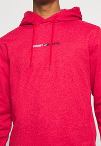 Sudadera con capucha roja de tela texturizada, con un logo frontal que dice "TOMMY JEANS" en blanco y negro. Incluye cordones en la capucha.