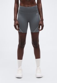 Αθλητικό άτομο που φοράει γκρι σορτς συμπίεσης Under Armour, λευκές κάλτσες μέσου μήκους και λευκά αθλητικά παπούτσια, στέκεται σε απλό φόντο.