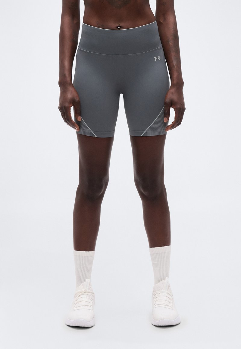 Αθλητικό άτομο που φοράει γκρι σορτς συμπίεσης Under Armour, λευκές κάλτσες μέσου μήκους και λευκά αθλητικά παπούτσια, στέκεται σε απλό φόντο.