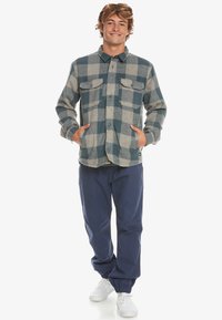 Flanellen overhemd in blauw en grijs geruit patroon met twee borstzakken, gecombineerd met marineblauwe broek en witte sneakers. Ontspannen pasvorm, met knoopsluiting.