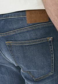 Tumed sinised denim teksad, millel on pruun nahkpatch ja "ONLY & SONS" tekst, nähtavad õmblused ja tagatasku disain.