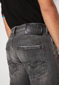 Jeans de mezclilla negros deslavados con un diseño de bolsillo trasero cosido, etiqueta de cinturón de cuero texturizado y un pequeño emblema rojo en el bolsillo.