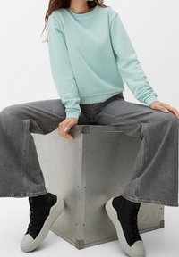 Mujer vestida con un sudadera azul claro, jeans grises de pierna ancha y zapatillas negras y blancas, sentada en una caja de metal con las piernas abiertas.