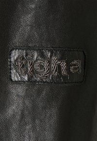 Svart läderpatch med präglad text. Slät textur med en lätt lyster; sömmarna framhäver kanterna och detaljerna i designen.