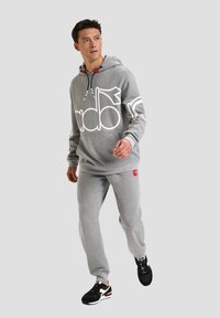 Felpa grigia con grande logo bianco, dettagli rossi e lacci. Pantaloni della tuta grigi con una piccola etichetta rossa. Sneakers nere con dettagli rossi e bianchi.