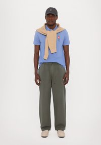 Light blue polo shirt with logo, beige sweater draped over shoulders, olive green trousers, and gray cap, paired with beige shoes.