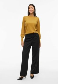 Maglione a maniche lunghe color senape con una vestibilità comoda, abbinato a pantaloni neri a gamba larga. L'outfit presenta una texture morbida e eleganti tacchi neri.