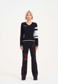 Black knit sweater with white striped sleeves and embroidered detail, paired with black flared jeans featuring red graphic embroidery.