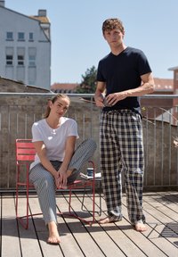 Dva lidé na dřevěné terase. Jeden má na sobě bílé tričko a pruhované pyžamové kalhoty, zatímco druhý má na sobě námořnické tričko a kostkované pyžamové kalhoty.