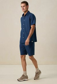 Korte mouwen marineblauw shirt met verticale strepen, gecombineerd met bijpassende shorts. Gedragen met beige sneakers en sokken. Zachte stoftextuur.