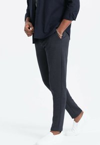Pantaloni slim-fit blu strutturati con tasche laterali, abbinati a scarpe bianche senza lacci e una camicia button-up scura.