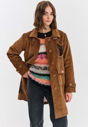 Brązowa kurtka z korduroy z guzikami, z przednią kieszenią i wyszywaną dekoracją, noszona na kolorowym, pasiastym swetrze z dzianiny.