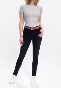 Kvinde iført en lys grå t-shirt med korte �ærmer, sorte skinny jeans, et farverigt vævet bælte og hvide sneakers, der står med den ene hånd i lommen.