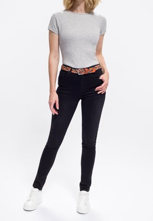 Femme portant un t-shirt gris clair à manches courtes, un jean skinny noir, une ceinture tissée colorée et des baskets blanches, se tenant avec une main dans la poche.