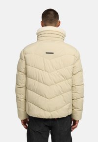 Pufasta jakna boje krem s visokim ovratnikom, naglašenim šavovima i logotipom na leđima, s teksturiranim završetkom.