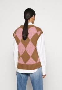 Chaleco de punto en argyle marrón y rosa con dobladillo acanalado, llevado sobre una camisa blanca con cuello, que presenta un corte holgado y mangas cortas.
