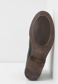 Chaussure noire avec une semelle brune texturée, présentant des rainures horizontales et un bout semi-arrondi. Détails de couture le long du bord et de la base.