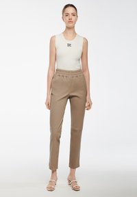 Canotta color crema con un piccolo logo nero; pantaloni beige tapered con vita elastica e cerniere laterali; il modello indossa sandali bianchi.