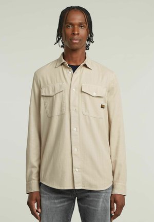 Beige Langarmshirt aus weichem Material mit zwei Vorderstaschen und abgerundetem Saum. Mit weißen Knöpfen und einem kleinen Logopatch.